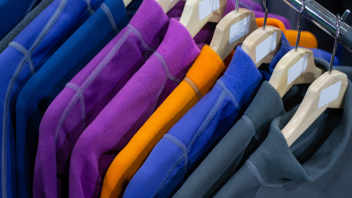 Fargerike fleecegensere i blått, lilla, oransje og grått henger på trekleshengere i en klesbutikk.
